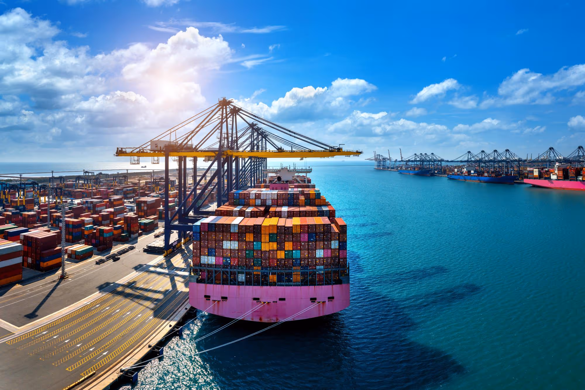 Large container ship docked at a busy port with colorful shipping containers and cranes under a partly cloudy sky.