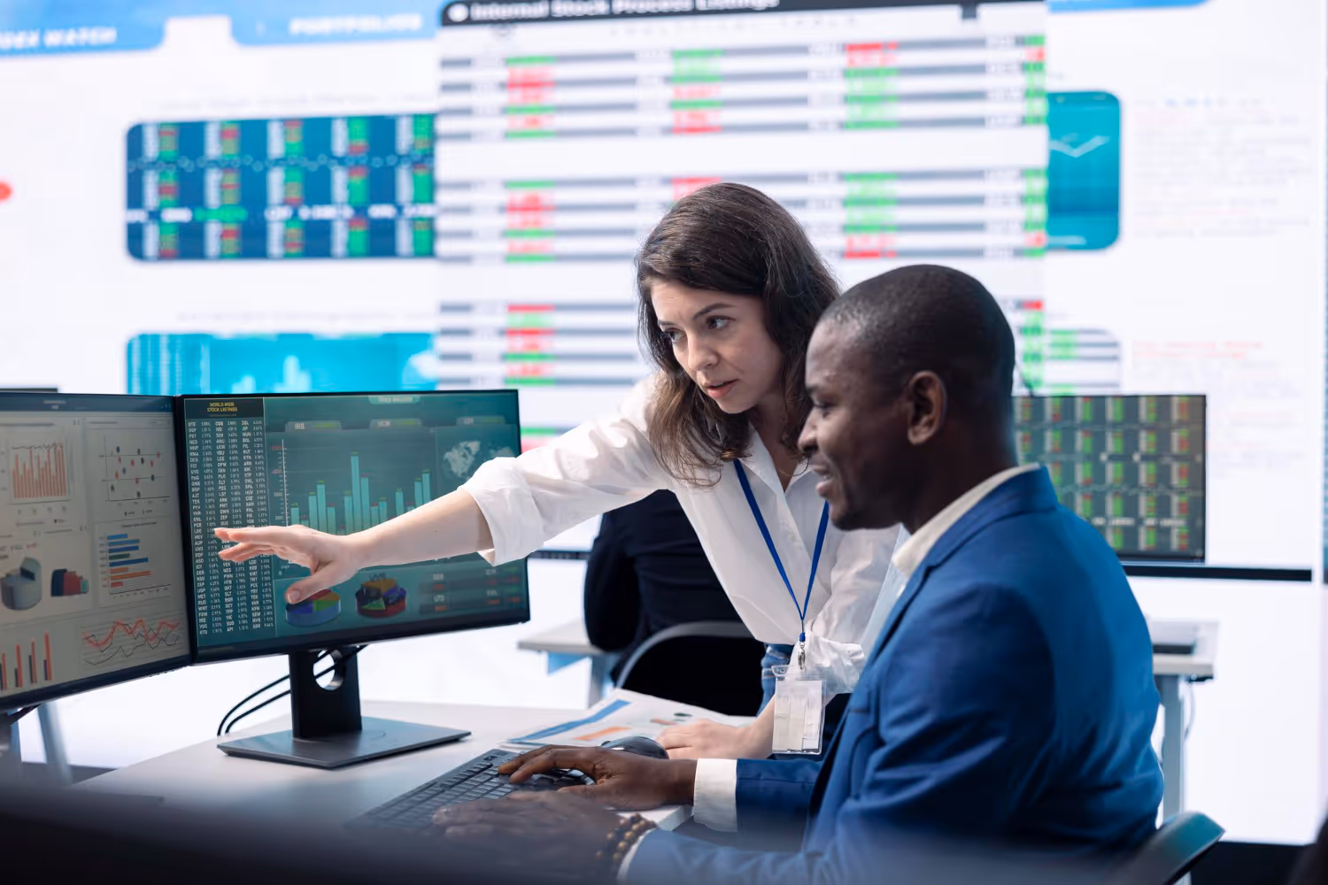 Two business professionals analyzing data charts and graphs displayed on dual computer monitors in a modern office.