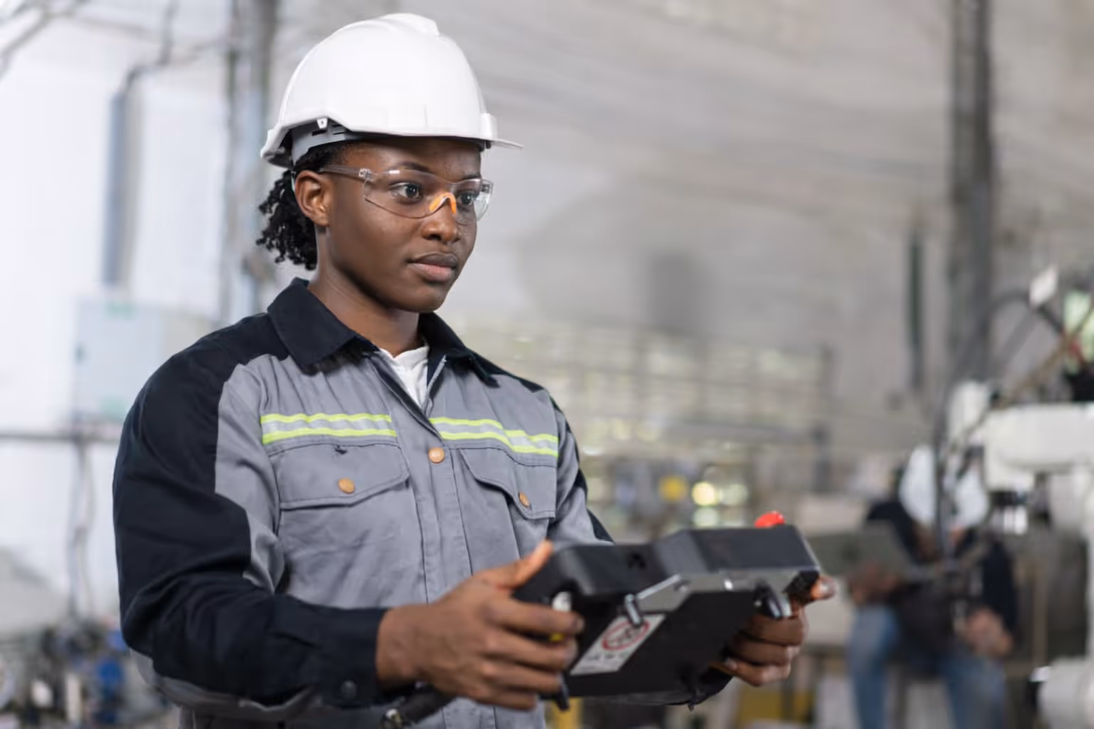 Worker in a hard hat and safety glasses using a digital controller in an industrial setting.