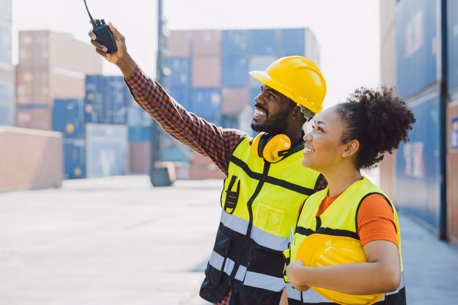 Two construction workers in reflective vests and hard hats at a shipping container yard, one holding a walkie-talkie and pointing.